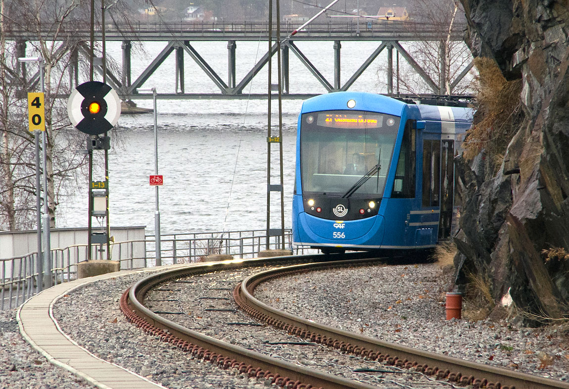 Bild: Ett tåg mot Gåshaga påbörjar stigningen upp till Baggeby från Herserud den 5 december 2015. I bakgrunden syns bron mellan fastlandet och Lidingö. Foto Markus Tellerup.