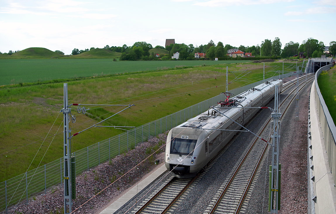 Ett tåg mot Stockholm bestående av X55 3345 vid Gamla Uppsala i juni 2017. I bakgrunden syns Kungshögarna, Gamla Uppsala kyrka och till höger mynningen till tunneln under området. Foto Markus Tellerup.