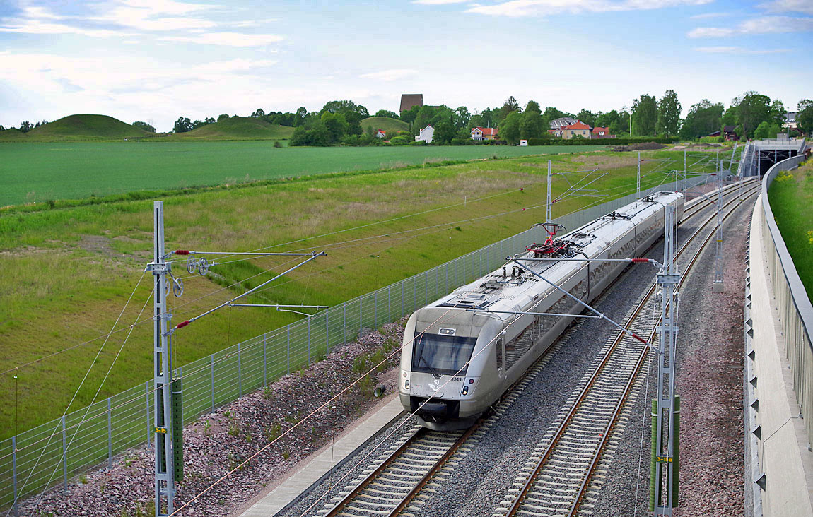 Ett tåg mot Stockholm bestående av X55 3345 vid Gamla Uppsala i juni 2017. I bakgrunden syns Kungshögarna, Gamla Uppsala kyrka och till höger mynningen till tunneln under området. Foto Markus Tellerup.