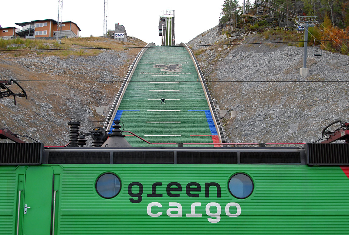 En backhoppare i Örnsköldsvik på väg att ta mark. Ett godståg mot Mellansel inväntar samtidigt avgång på nuvarande Örnsköldsvik C som delvis är byggd på en viadukt över hoppbackens nedre del. Foto 2010, Markus Tellerup.