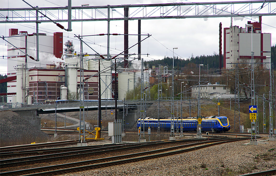 X62 001 som tåg Sundsvall-Umeå anländer till Timrå. I bakgrunden Östrands massafabrik. Ådalsbanans moderna signalsystem ERTMS saknar ljussignaler men har tavlor (gul pil på blå botten) som markerar signalernas position. Foto 2015, Markus Tellerup.