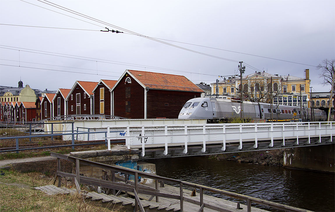 Ett södergående snabbtåg med X2 2015 främst passerar sjöbodarna i Hudiksvall strax före stationen. Till höger syns tunneln genom Stadshotellet. Foto i april 2015, Markus Tellerup.